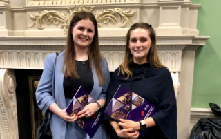 Áine Fealy and Caoimhe Newell in Houses of the Oireachtas standing in front of a decorative fireplace, Irish flags and a portrait of the president.