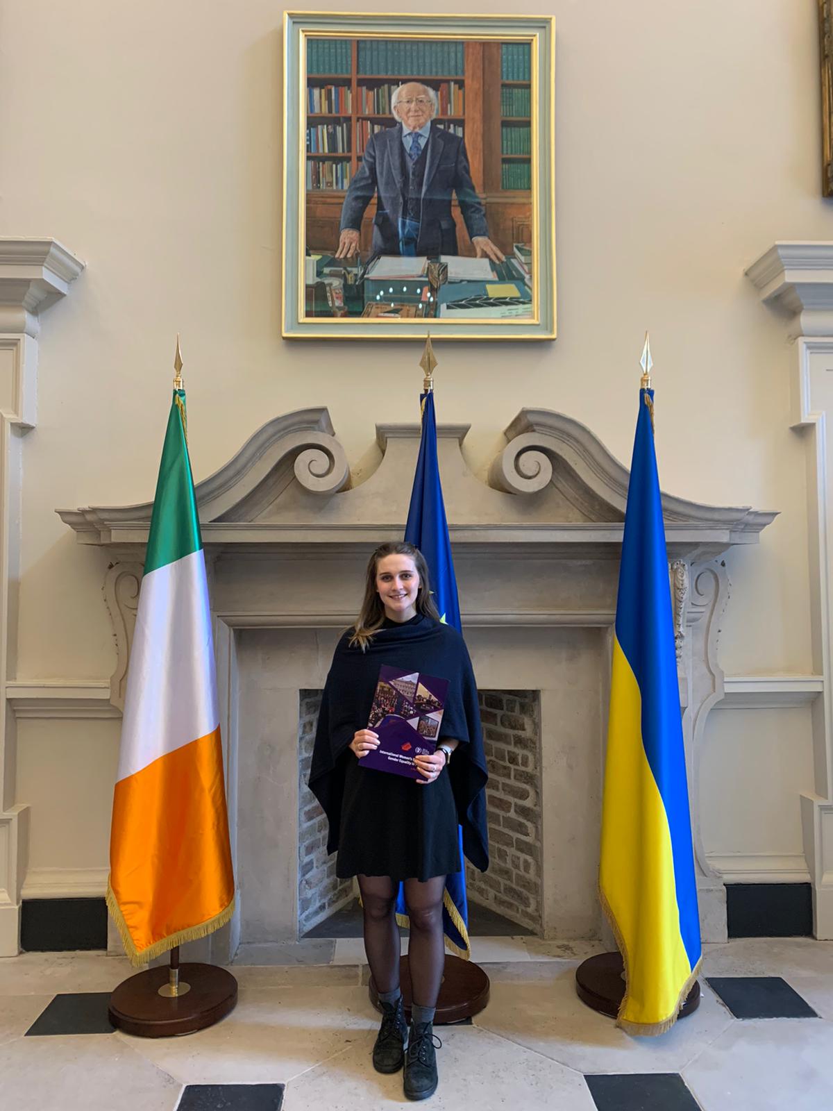 Caoimhe Newell in Houses of the Oireachtas standing in front of a decorative fireplace, Irish flags and a portrait of the president.