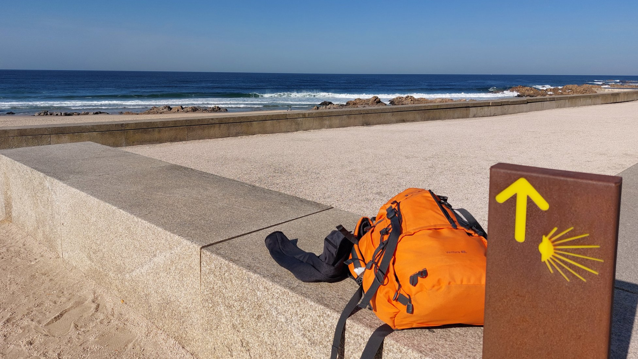 The portugal coastline with a bright orange hiking backpack laying on a stone wall.